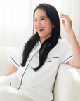 Woman in a white top with navy logo sitting on a couch, smiling.