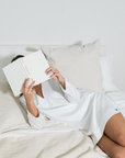 Person lying on a bed reading a book in a white dress.