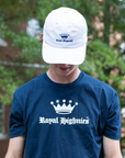Person wearing a navy 'Royal Highness' t-shirt and white cap with logo, outdoors.