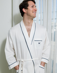 Man wearing a white robe with navy trim and logo standing in a room with light-colored walls and a window.