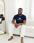 Man sitting on a white couch wearing a navy blue t-shirt with a logo and white pants with navy logo in a bright room.