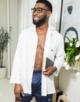 Man wearing a white long sleeve shirt over navy shorts holding a book in a room with decors.