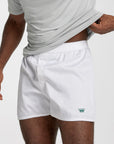 White boxer shorts with an elastic waistband and a small green crown logo on the lower left leg, shown on a model wearing a light gray t-shirt against a plain light gray background.