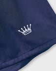 Close-up of a navy blue boxer shorts with a small white embroidered crown logo on the left side, shown against a light neutral background.