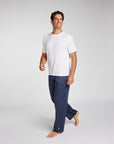 A man wearing a white short-sleeve crewneck t-shirt and navy blue pajama pants, walking barefoot against a light gray studio background.