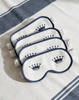 Four white sleep masks with navy blue borders and crown designs, stacked diagonally on a white and navy striped blanket.