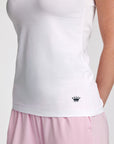 Close-up of a person wearing a white tank top with a small navy crown logo near the lower front, paired with light pink bottoms, against a plain light background.