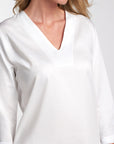 A close-up of a woman wearing a white long-sleeve blouse with a V-neckline, shown against a light gray studio background.
