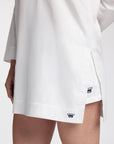 Close-up of a white tunic with a side slit, worn by a model. The hem features a small navy blue embroidered crown logo
