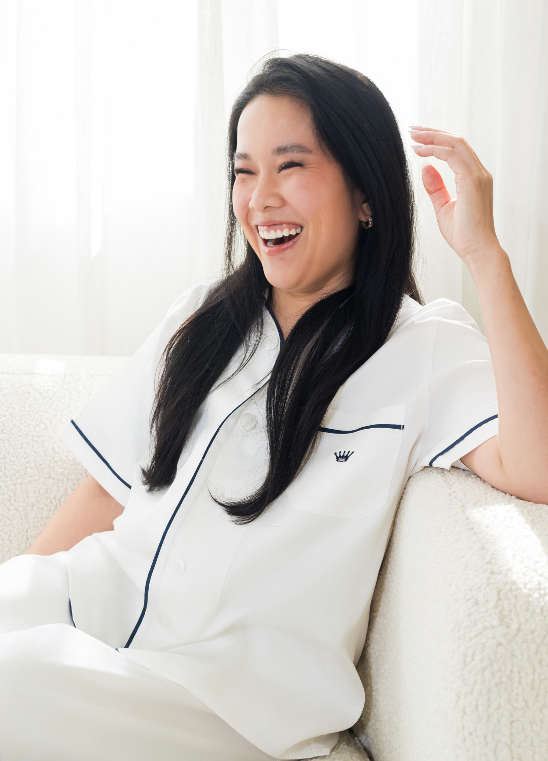 Woman in a white top with navy logo sitting on a couch, smiling.