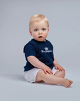 Baby sitting on a plain background wearing a navy blue shirt with white text and white shorts.