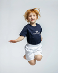 Child wearing a navy blue t-shirt with a logo and white shorts on a light gray background
