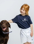 Child wearing a navy blue t-shirt with a logo, standing next to a brown dog on a light gray background