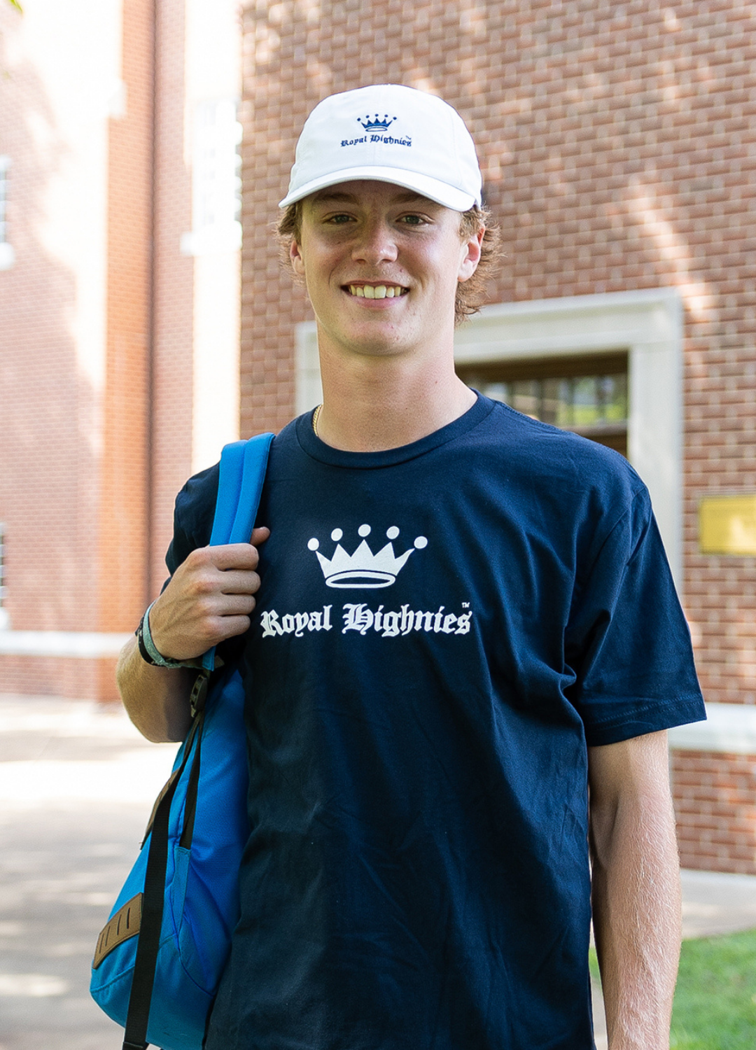 Person wearing a navy blue 'Royal Highness' t-shirt with a white cap, standing outdoors.
