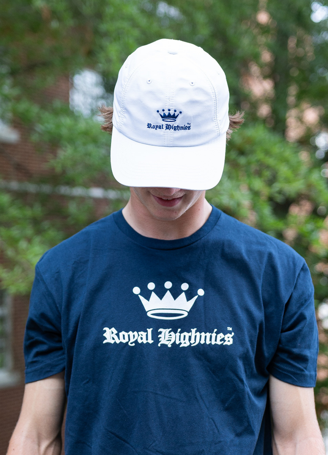 Person wearing a navy 'Royal Highness' t-shirt and white cap with logo, outdoors.