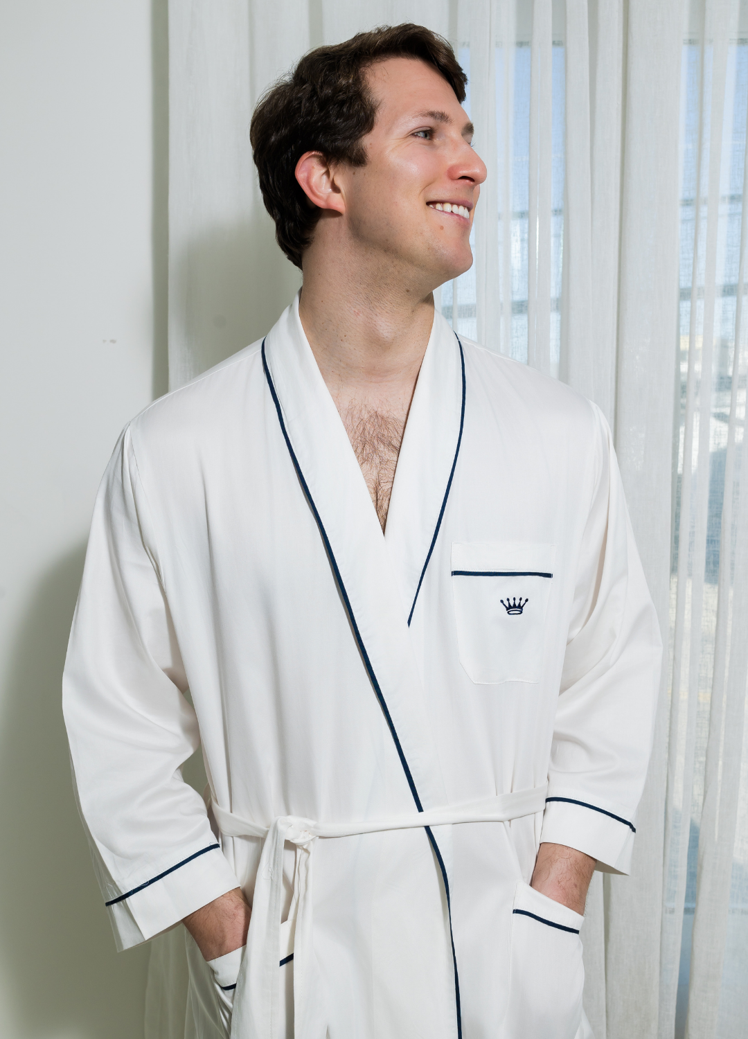 Man wearing a white robe with navy trim and logo standing in a room with light-colored walls and a window.