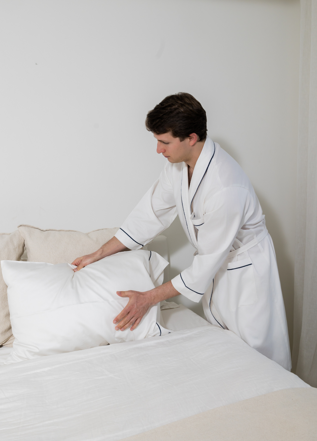 Person in a white robe adjusting pillows on a bed in a minimalistic room.