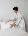 Person in a white robe adjusting pillows on a bed in a minimalistic room.