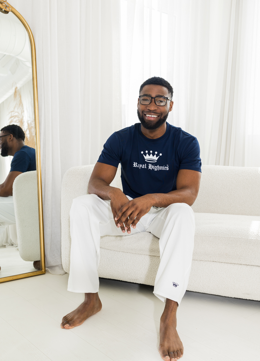Man sitting on a white couch wearing a navy blue t-shirt with a logo and white pants with navy logo in a bright room.
