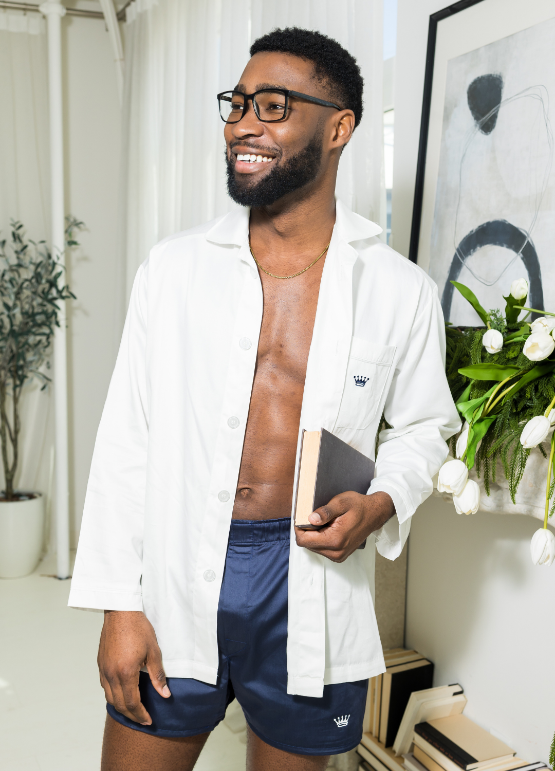 Man wearing a white long sleeve shirt over navy shorts holding a book in a room with decors.