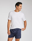 A man wearing a white short-sleeve athletic t-shirt and navy blue boxer shorts, standing against a light gray studio background.