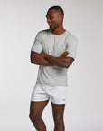 A man wearing a light gray short-sleeve athletic t-shirt and white boxer shorts, standing with arms crossed against a light gray studio background.