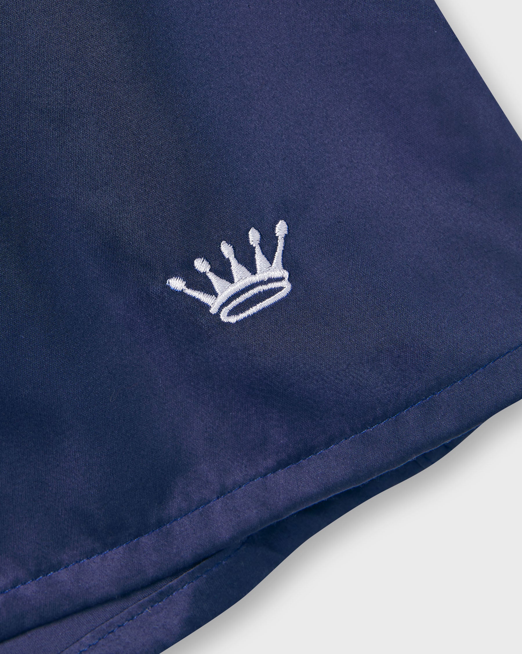 Close-up of a navy blue lounge pants with a small white embroidered crown logo on the left side, shown against a light neutral background.