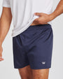 A close-up of a person wearing navy boxer shorts with a small white crown logo near the lower left leg, paired with a white t-shirt against a light gray studio background.