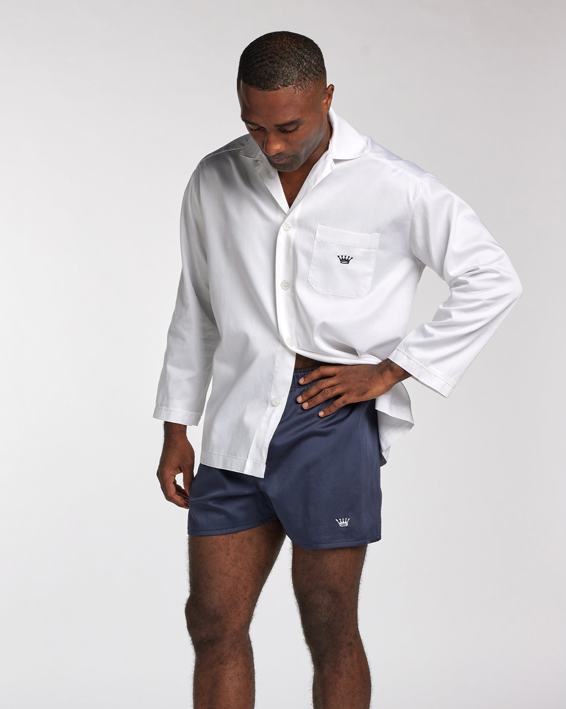 A man wearing a white long-sleeve button-up shirt with a chest pocket and navy blue shorts, standing against a light gray studio background.