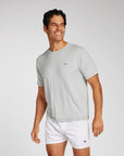A man wearing a light gray short-sleeve athletic t-shirt and white boxer shorts, standing against a light gray studio background.