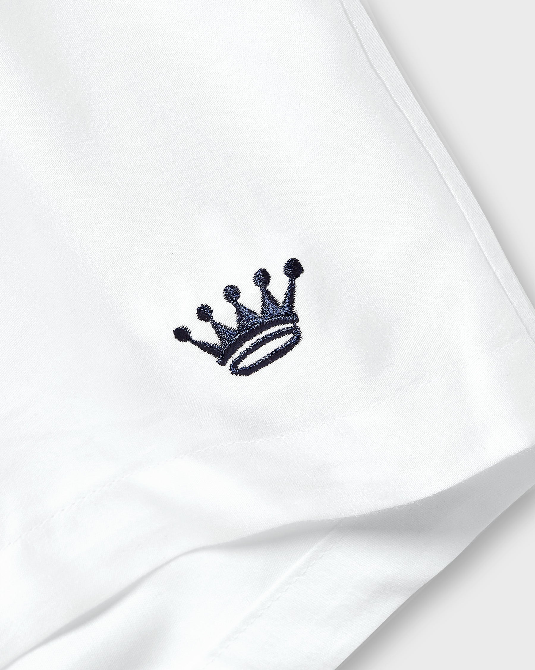 A close-up of a white fabric item, likely a shirt or garment, featuring a small navy embroidered crown logo on the chest.