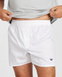 White boxer shorts with an elastic waistband and a small navy crown logo on the lower left hem, shown on a model lifting a light gray shirt.