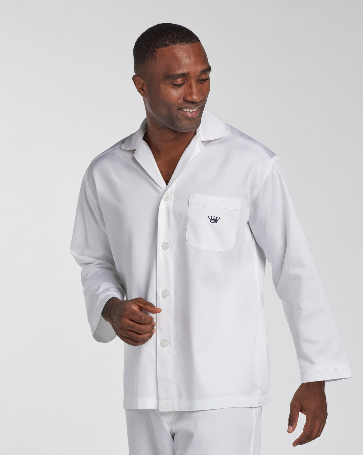 A man wearing a white long-sleeve button-front pajama top with a chest pocket and a small embroidered crown logo, photographed against a light gray studio background.