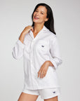 A woman modeling a white long-sleeve button-up shirt and matching shorts set, both featuring a small navy crown logo on the chest and hem, against a light gray studio background.