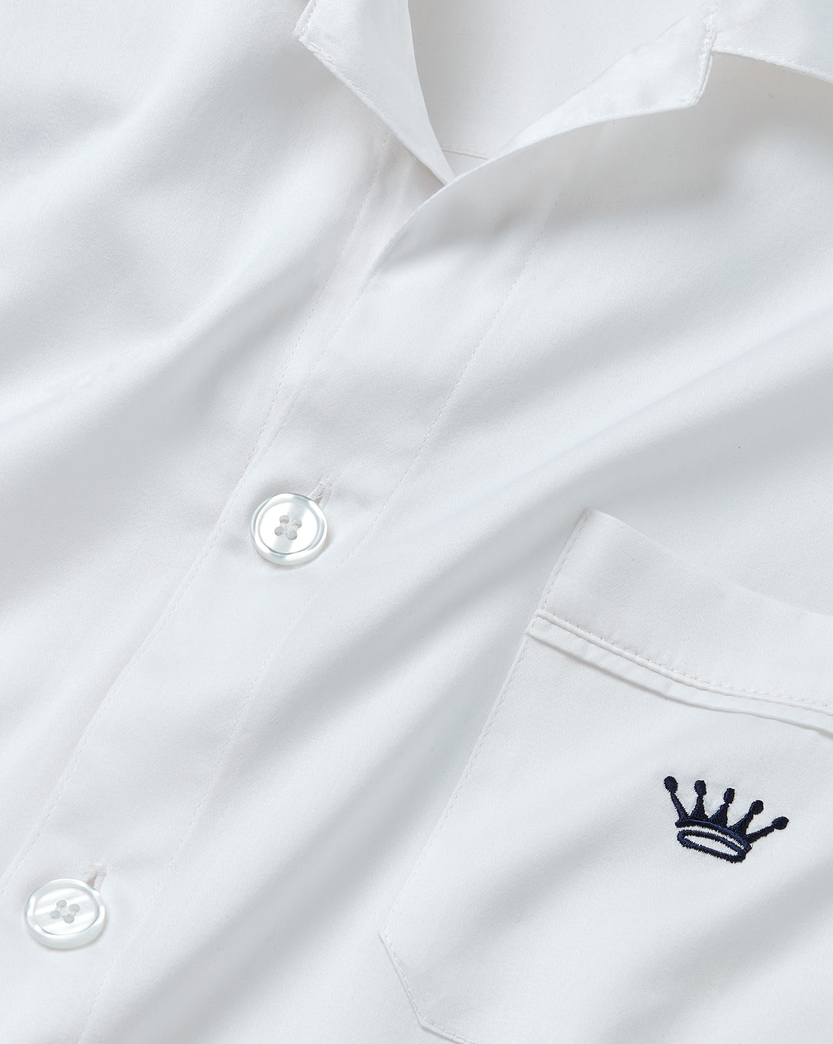 Close-up of a white long-sleeve button-up shirt with a chest pocket featuring a small navy embroidered crown logo.