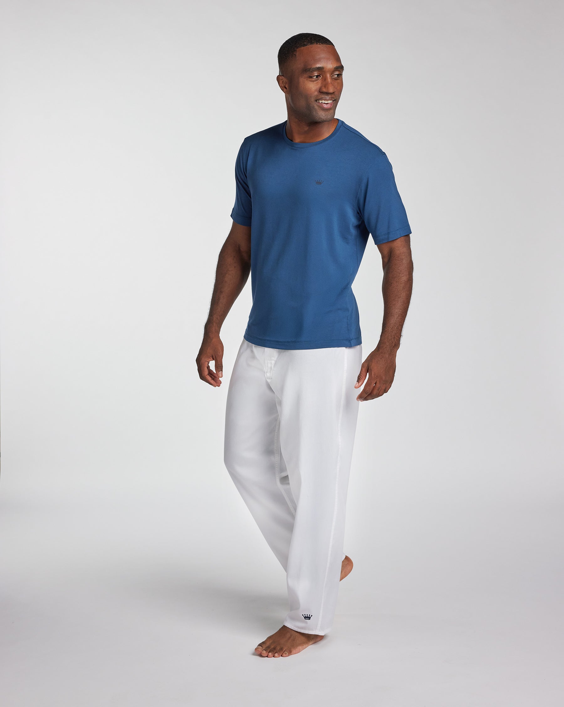 A man wearing a blue short-sleeve crewneck t-shirt and white pants, walking barefoot against a light gray studio background.