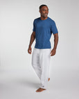 A man wearing a blue short-sleeve crewneck t-shirt and white pants, walking barefoot against a light gray studio background.