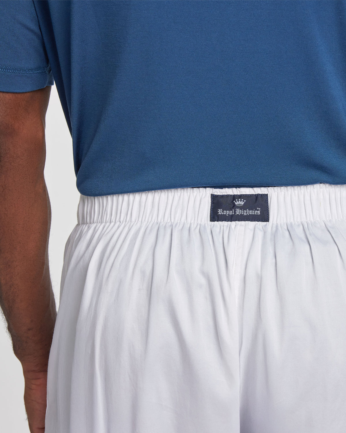 Close-up of a person wearing white lounge pants with a black elastic waistband and a visible Royal Highnies label, paired with a blue shirt against a light gray background.