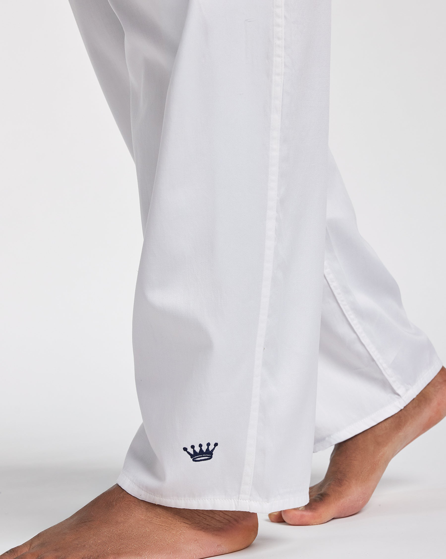 White wide-leg pants shown on a model from the calves down, featuring a small navy blue embroidered crown logo near the hem and a vertical seam running down the side.