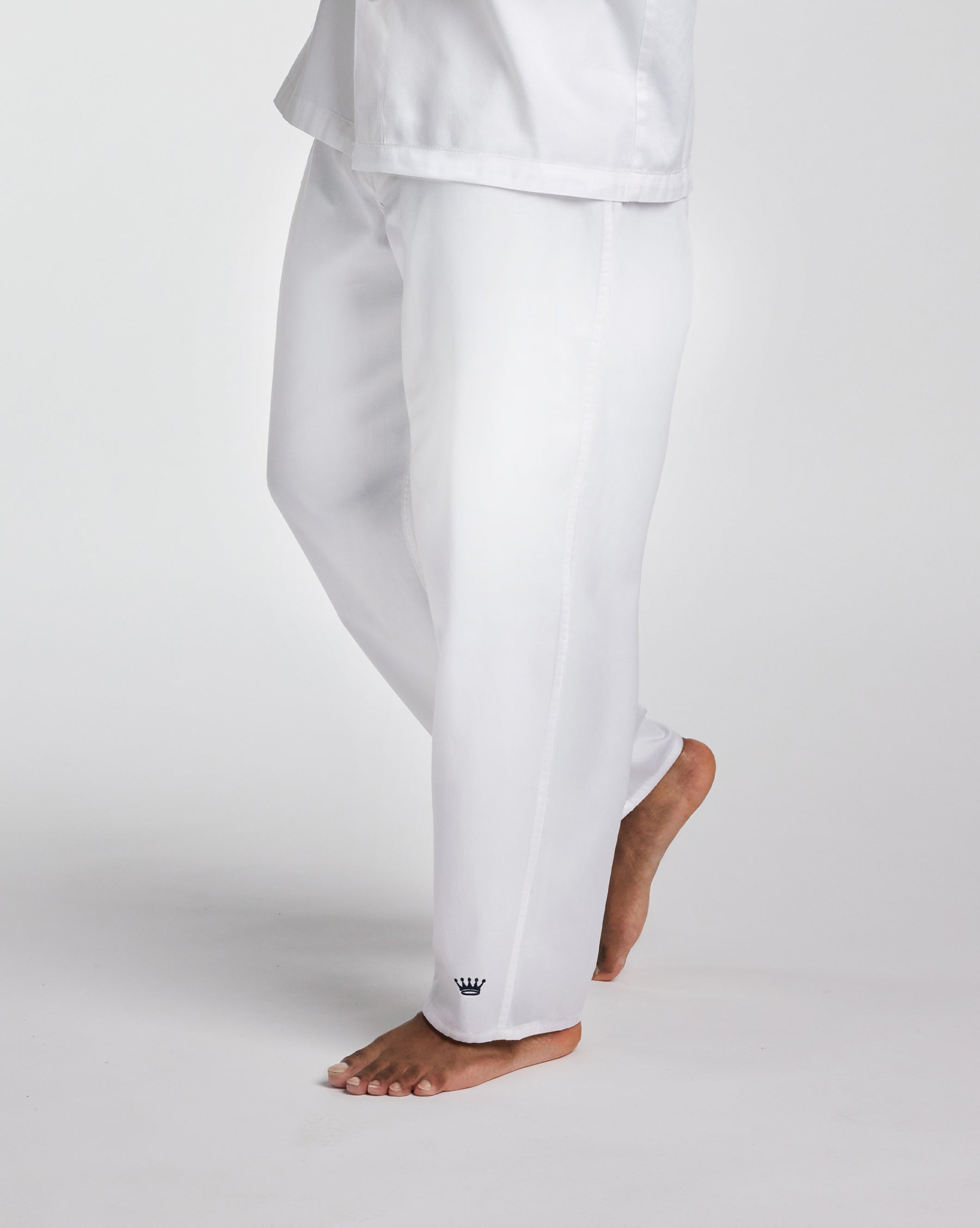 White lounge pants worn by a model, shown from the waist down in a side profile against a light gray studio background. A small navy crown logo is visible near the ankle.