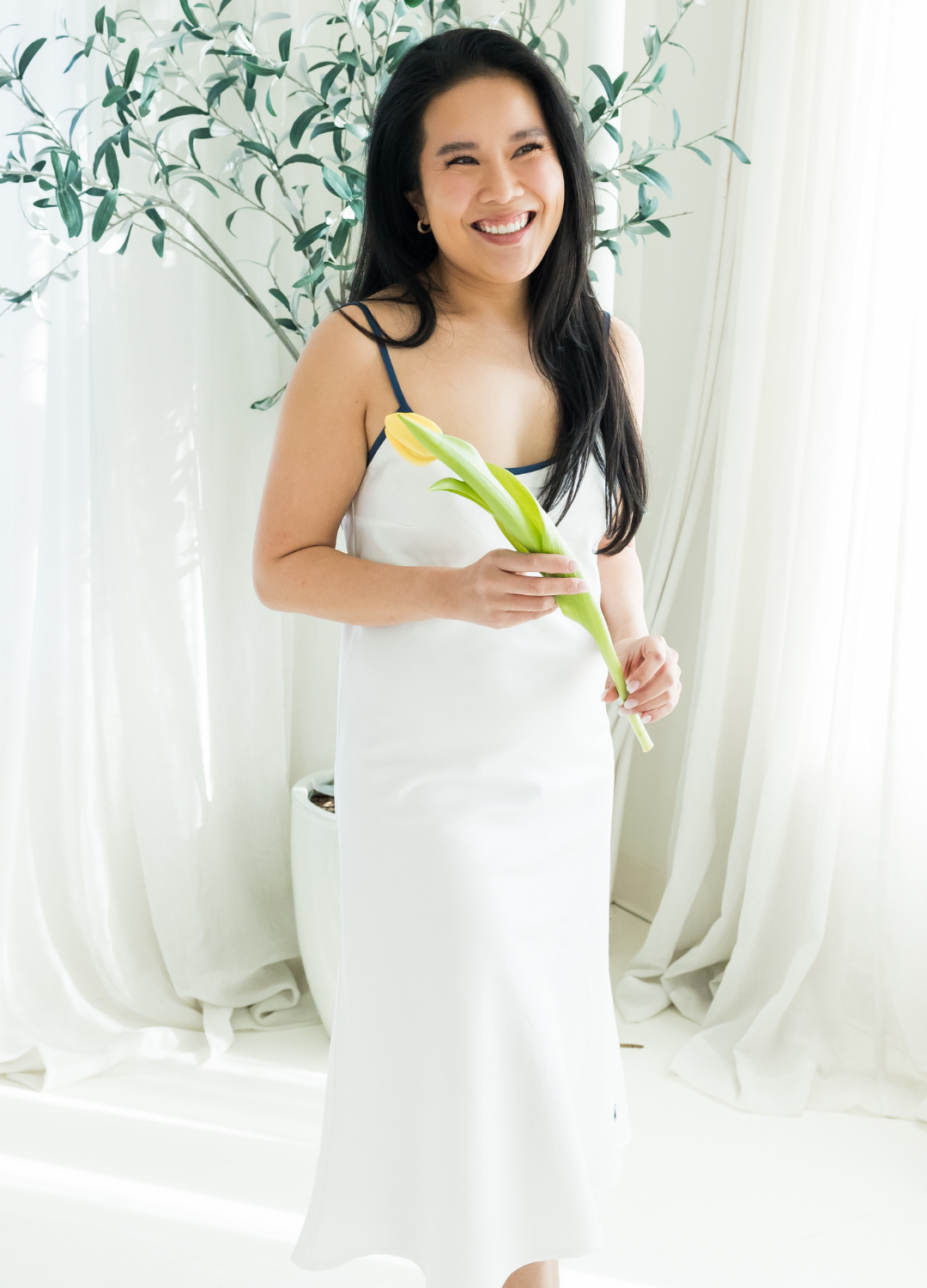 Woman in a white night gown holding a flower in front of white curtains