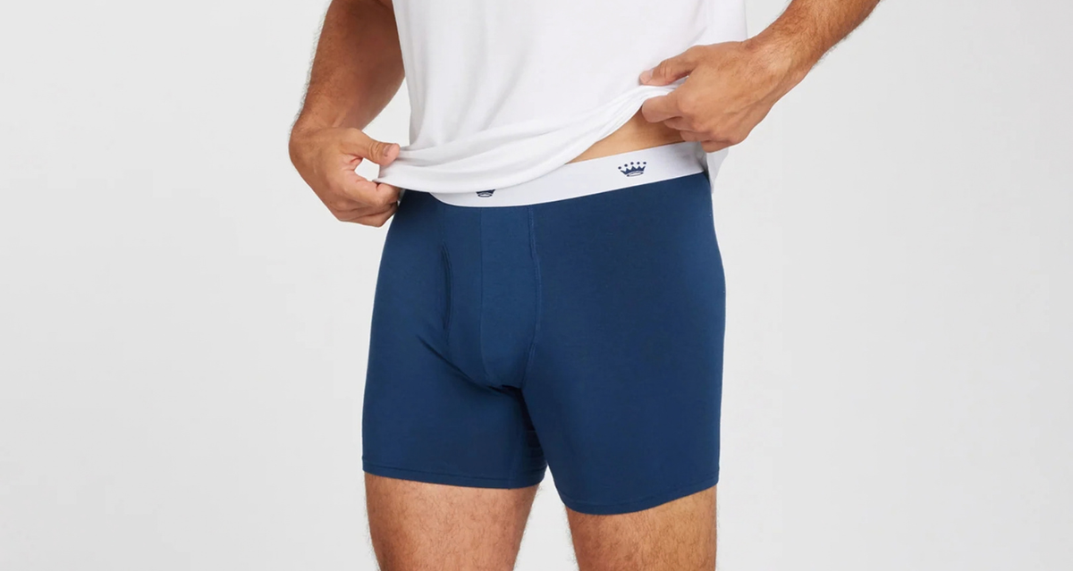 A man wearing navy blue boxer briefs and lifting the hem of a white t-shirt to show the elastic waistband, against a light gray background.