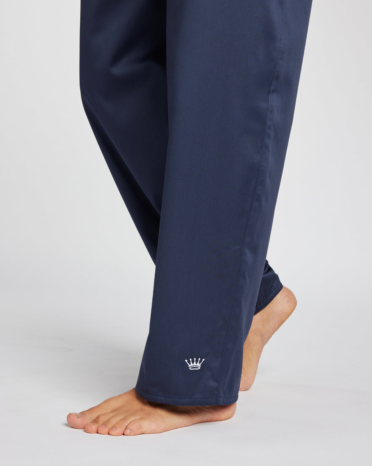 Close-up of a person wearing navy blue pants with a small white crown logo near the cuff, photographed against a light gray studio background.