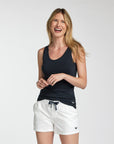 A woman smiling and wearing a dark navy tank top and white shorts with a drawstring, photographed against a light gray studio background.