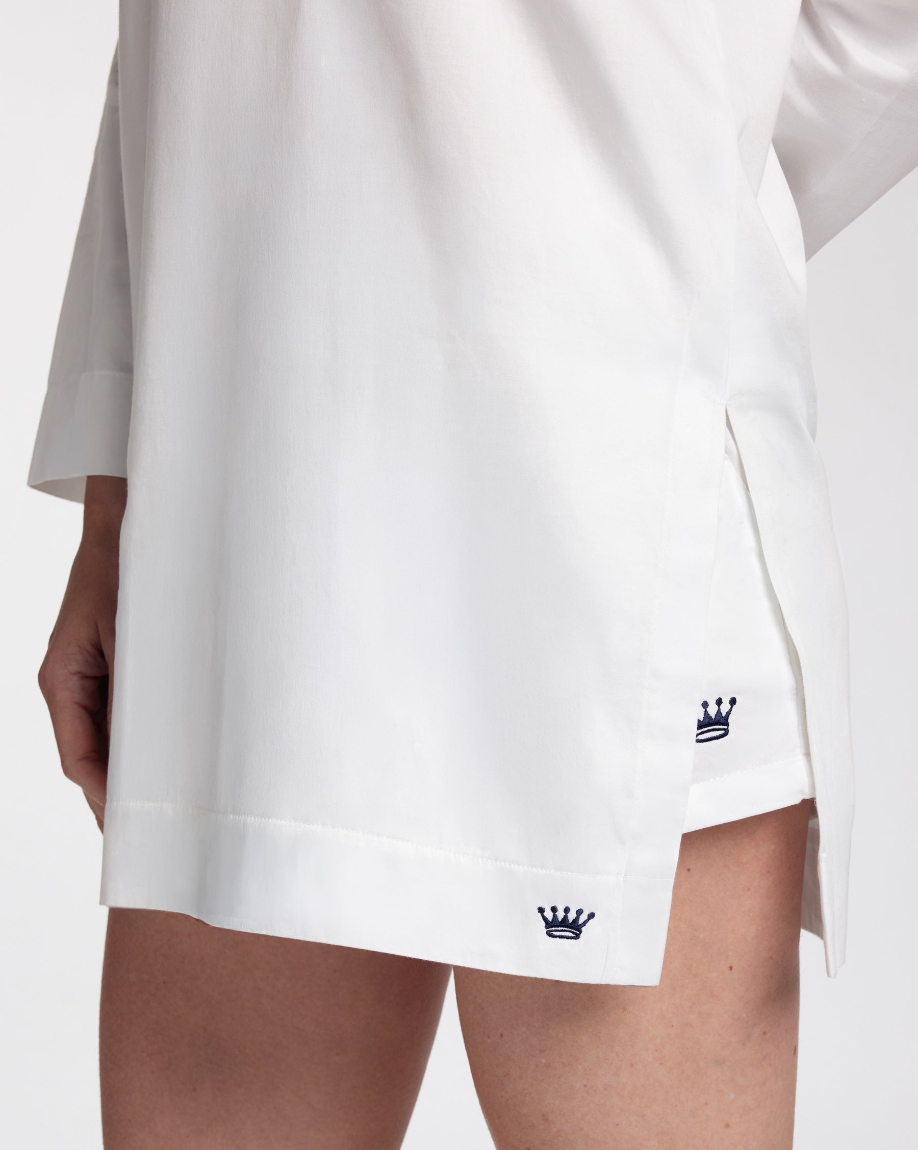 Close-up of a white tunic with a side slit, worn by a model. The hem features a small navy blue embroidered crown logo