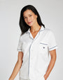 A woman wearing a white short-sleeve pajama top with thin dark piping and a small embroidered crown logo on the chest pocket, photographed against a light gray studio background.